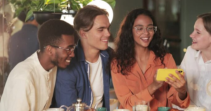 Attractive girl holding phone horizontally while showing friends funny photos. Millennial cheerful people laughing and looking at mobilephone screen while sitting in cosy cafe.