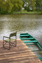 An empty chair and a fishing rod on a wooden pier by the lake, a green boat near the pier. Outdoor recreation.
