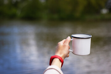 The girl holds in her hands a metal cup of coffee near the lake. Close-up.