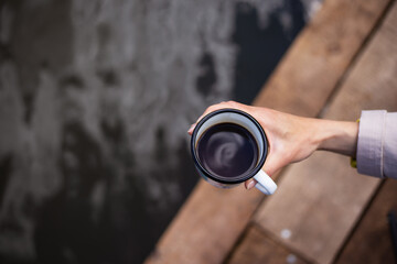 A girl holds a metal cup of coffee in her hands near the lake while sitting on a wooden pier. Close-up, top view.