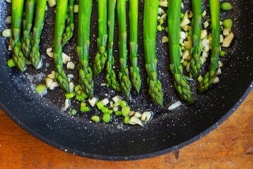 Tasty green asparagus is fried in a pan with garlic, next to it is a blue striped towel. Close-up, top view.