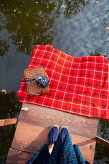 The old camera lies on a suitcase on a red plaid. Wooden bridge by the lake. Legs of the girl in the frame.