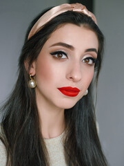 Close up headshot portrait of young natural looking casual beautiful brunette woman with trendy makeup and vibrant red lips in hair hoop and pearl earrings domestic room background