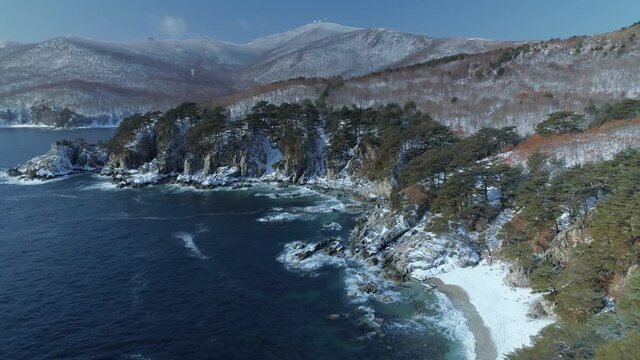 Aerial forward rocky seashore, blue ocean scenic coastline landscape, strong waves hitting rocks. Winter snowy forest, high hills. Famous Vityaz bay, Vladivostok Russia natural landmark. Sunny day. 4k