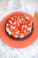 Strawberry cheesecake decorated with heart-shaped strawberries lies on a coral-colored plate, stands on a napkin and a wooden table. Illustration of a cheesecake recipe with strawberries.