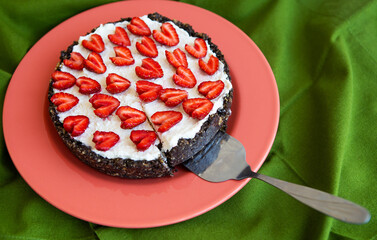 A strawberry cheesecake decorated with heart-shaped strawberries lies on a coral-colored plate, stands on a green napkin, cut off a slice. View from above.