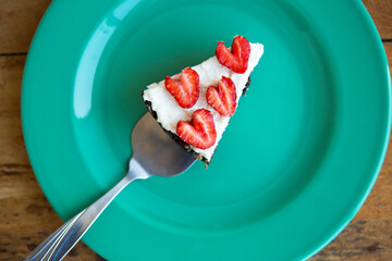 A piece of strawberry cheesecake decorated with heart-shaped strawberries lies on a mint-colored plate and stands on a checkered napkin. View from above.