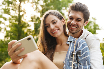 Image of young happy couple smiling and holding cellphone in park