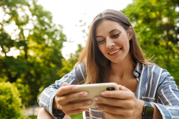 Image of young woman smiling and holding cellphone in park