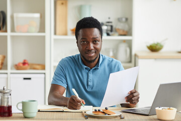 Obraz na płótnie Canvas Portrait of African young man planning his day and making notes in note pad while sitting at the table in the kitchen
