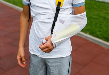 Teenager with broken arm next to sports ground. Boy in white t shirt with hand splint in a arm sling for support and reducing pressure on neck. Injury, traumas, accident by kids
