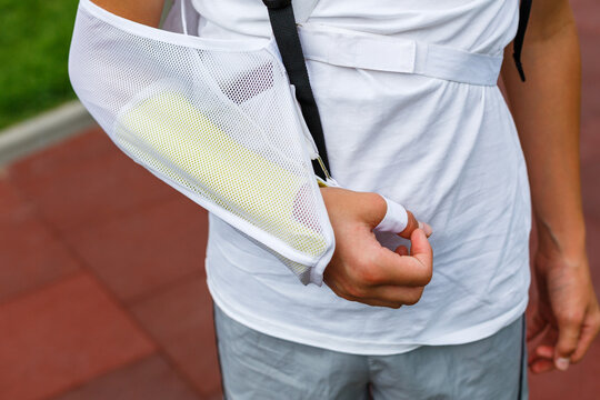 Teenager with broken arm next to sports ground. Boy in white t shirt with hand splint in a arm sling for support and reducing pressure on neck. Injury, traumas, accident by kids