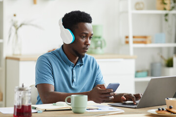 African man in headphones concentrating on his online work he using laptop and mobile phone while working at home