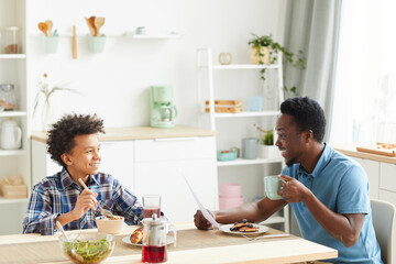 Fototapeta na wymiar African father talking to his son while they sitting at the table and having breakfast in the kitchen at home
