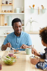 African young father has breakfast together with his son at the table in the kitchen they smiling and talking