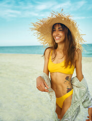 Beautiful smiling latin woman in bright yellow bikini and straw hat walking on beach by ocean seashore. Stylish girl enjoying summer tropical vacation at sea.