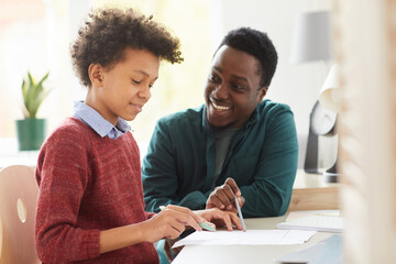 African smiling teacher teaching his student he pointing at paper and explaining him the new material at the desk at home