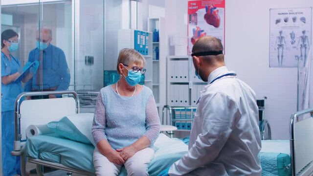 Physician discussing lungs diagnostic of retired old woman who sits on hospital bed in modern private clinic during COVID-19 pandemic healthcare global crisis. Doctor in protective equipment from