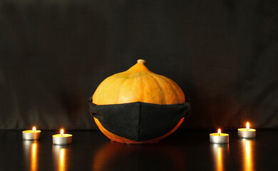 Halloween pumpkin in a black protective mask next to burning candles on a black background. Concept of preparation for the celebration of Halloween in the conditions of coronavirus. Copy space