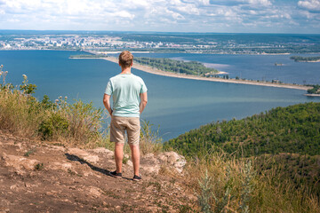 A young blond man stands with his back to the camera high on a mountain overlooking a Grand panorama of the Volga river and the Zhiguli mountains. Russian tourism