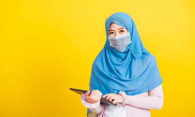 Asian Muslim Arab, Portrait of happy beautiful young woman Islam religious wear veil hijab and face mask protect she quarantines disease coronavirus touch on tablet screen isolated yellow background