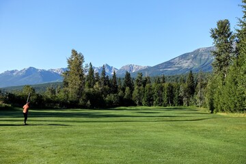 A beautiful view of a golf course on a sunny summer morning with the sun shining and blue sky, surrounded by tall trees.  Golf in nature in British Columbia, Canada.  A woman swinging club on fairway