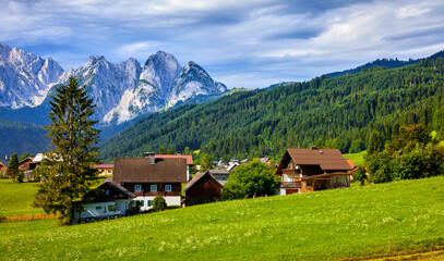 Austrian Alps High Mountains with high snowy tops. Alpine Village in forest and lawns with green grass. Traditional alpine village houses on the fields. Sunny morning