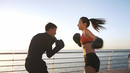 Young female in sportswear is boxing with her skillful coach. They are dodging punches while training on promenade by sea - Powered by Adobe