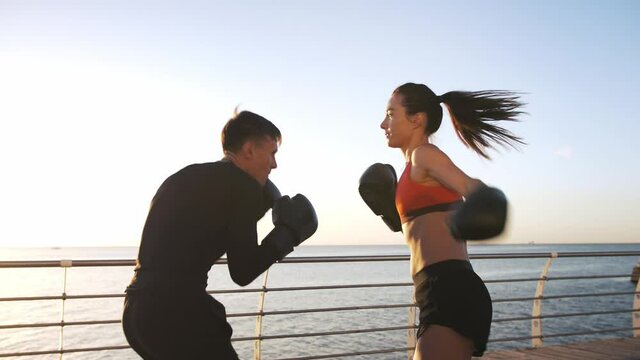 Young female in sportswear is boxing with her skillful coach. They are dodging punches while training on promenade by sea