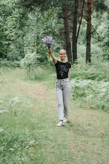 young woman with a tail of hair in a black t-shirt and gray jeans in the woods holding blue wildflowers in her hands