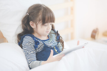 Cute asian little girl enjoy watching cartoon on smart tablet while sitting on bed in kid's bedroom at home.Photo series of family, kids and happy people concept