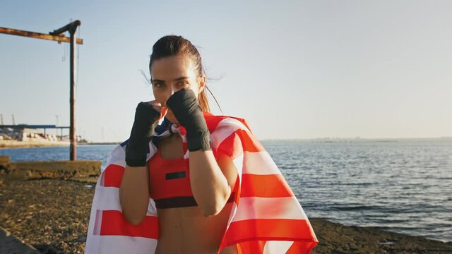 Young woman in sportswear, wrapped in flag of USA. She is boxing by hands in black bandage tape. Training by seaside, near city port. Slow motion