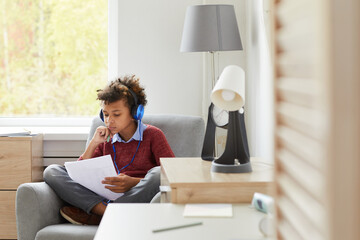 Fototapeta premium African boy in headphones sitting on armchair with papers he learning lessons or preparing for the exam at home