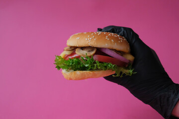 Hand in black glove holding fresh burger with mushrooms, tomatos, lettuce, onion and cheese on bright pink background.
