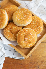 Freshly baked hamburger buns lie on a wooden tray on the table with a white napkin. Yellow flavored pastries with white sesame seeds.