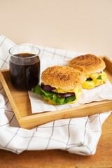 Large and tasty grilled hamburgers with sesame seeds lie on a wooden tray with a glass of cola. Wooden table and white checkered napkin. Close-up.