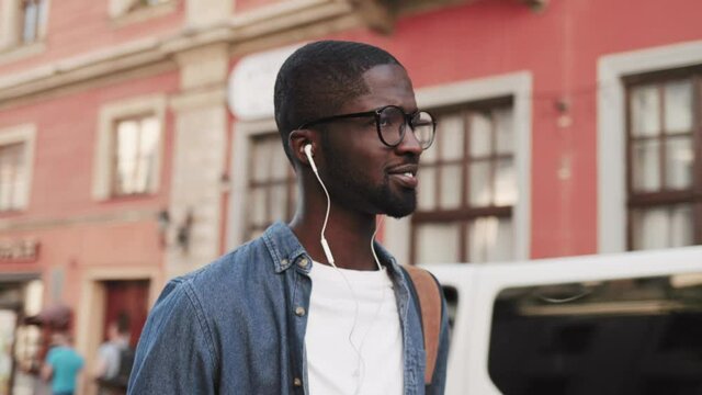 Handsome African American guy walking in city and listening music with headphones. Happy smiling man enjoying music.
