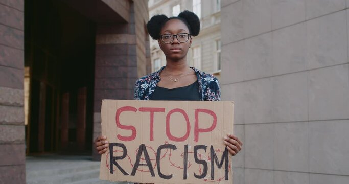 Portrait of afro american girl holding in her hands stop racism phrase cardboard. Female activist supporting anti racism campaign at city street. Concept of equal human rights
