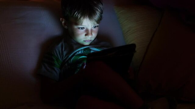 Dim light scene of a child sitting on the couch playing with a tablet. The light from the display illuminates the child’s face and creating an evocative atmosphere with light and color effects. 