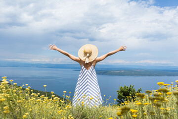Rear view of young woman wearing striped summer dress and straw hat standing in super bloom of wildflowers, relaxing with hands up to the sky, enjoing beautiful view of Adriatic sea nature, Croatia.