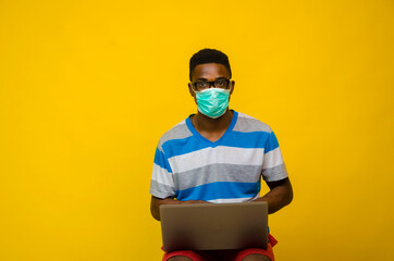 a young african man wearing face mask isolated over yellow background prevent, prevented himself from the outbreak in the society while operating his laptop