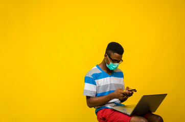 young handsome african man wearing face mask isolated over yellow background preventing himself from the outbreak in the society operating his phone while his laptop is on his leg