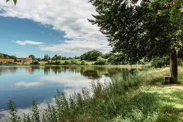 Obraz premium A calm and tranquil zen-like sunny day by a lake with reflections on the water.Park with green grass and trees.Tranquil Background.Colorful scenery.Dream relax concept.Picturesque view of nature