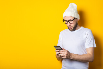 Bearded young man in a hat and glasses looks in surprise at the phone on a yellow background.