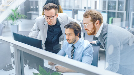 In Bright Modern Office: Young Businessman Sitting and Working at His Desktop Computer with Project Manager and Team Leader Standing Beside Him, Have Discussion, Finding Problem Solution 