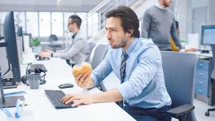 In the Bright Modern Office Young Businessman Eats Hamburger Sitting at His Desktop Computer, He Chewing the Bun and Continues to Work During His Lunch. In the Background Colleagues Working
