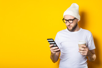 Bearded young man with cardboard glass with coffee looking at phone on yellow background.