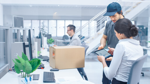 Happy Delivery Man in Corporate Office, Hands Package to a Beautiful Businesswoman, She signs Her Signature on the Tablet Computer. Big Bright Modern Business Company Office with Professionals