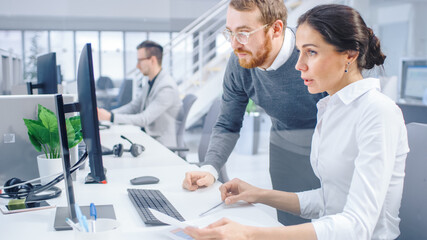 Beautiful Businesswoman Uses Desktop Computer, Consults Her Project Manager about Documents with Graphs and Statistics. In the Background Big Corporate Firm Office