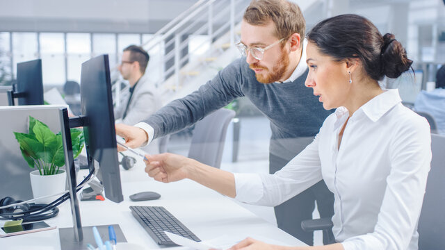 Beautiful Businesswoman Uses Desktop Computer, Consults Her Project Manager about Documents with Graphs and Statistics. In the Background Big Corporate Firm Office